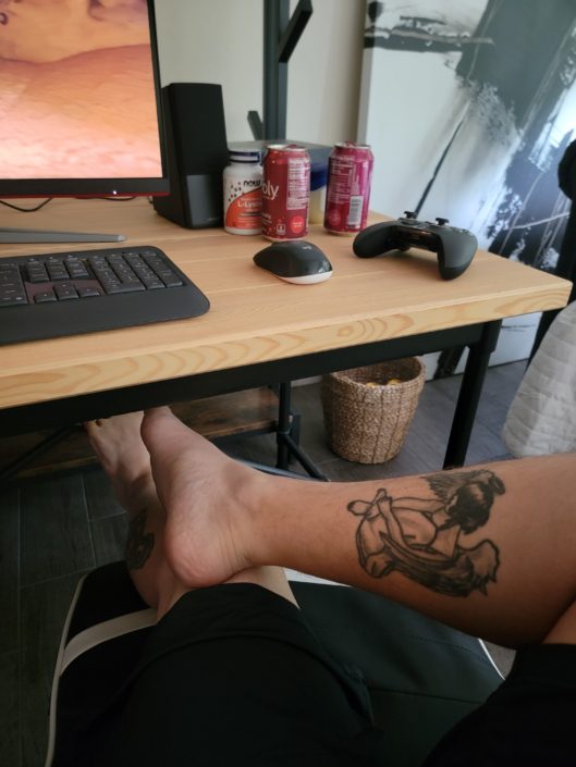 A person with tattoos on their feet is sitting at the computer desk.